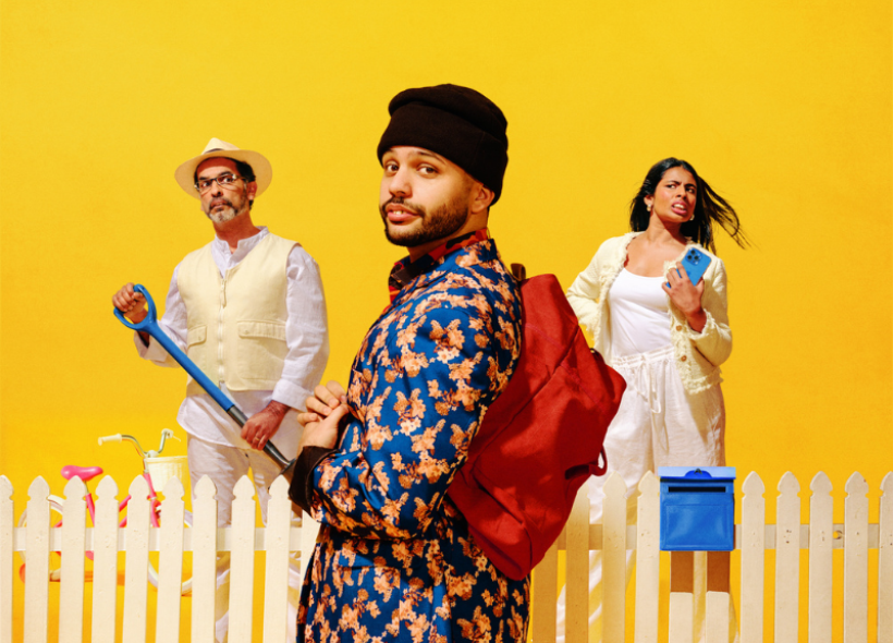 Three people stand in front of a white picket fence with a bright yellow background. One man wears floral print and a beanie, while the others wear light clothing and hold garden tools.