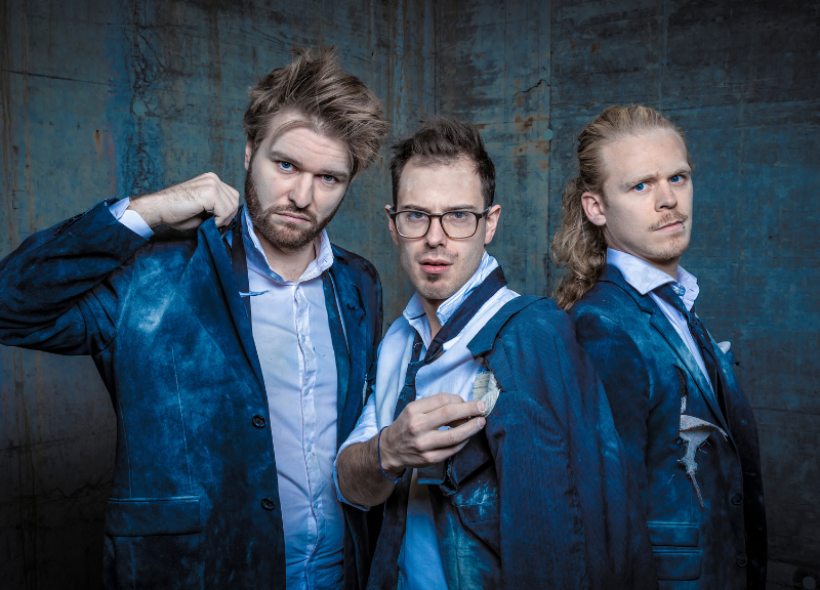 Three band members posing wearing blue suits on a blue distressed backdrop.