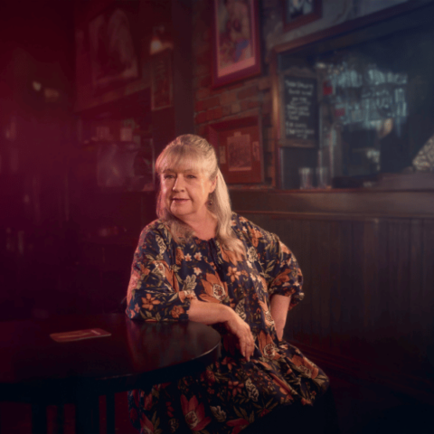 An older woman with long gray hair sits at a dark café table, wearing a floral dress. Warm light highlights her face.