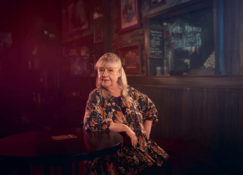 An older woman with long gray hair sits at a dark café table, wearing a floral dress. Warm light highlights her face.