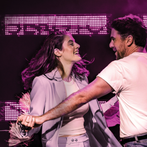 A woman and a man dance together on stage under purple lighting, both smiling. The man wears a white t-shirt and jeans, and the woman wears a light-colored suit.