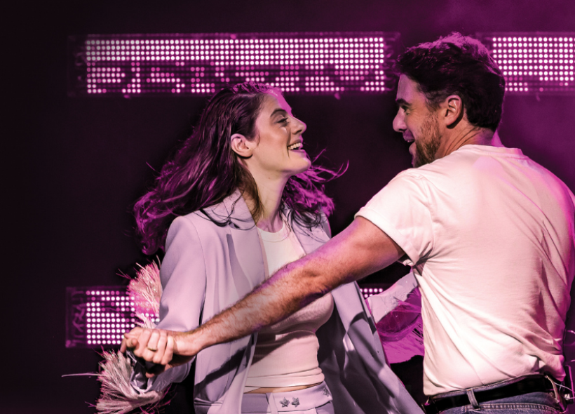 A woman and a man dance together on stage under purple lighting, both smiling. The man wears a white t-shirt and jeans, and the woman wears a light-colored suit.