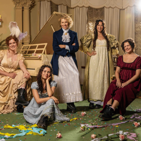 Five women in Regency-era dresses pose playfully in an elegant room