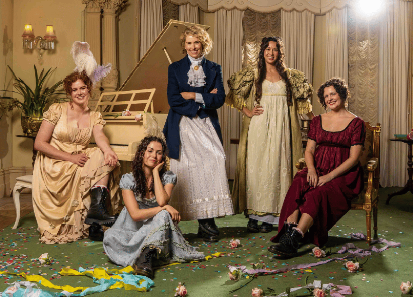 Five women in Regency-era dresses pose playfully in an elegant room