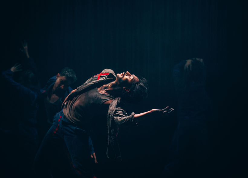 A dancer dramatically poses on stage under moody lighting with a dark background. Other dancers are partially visible. 