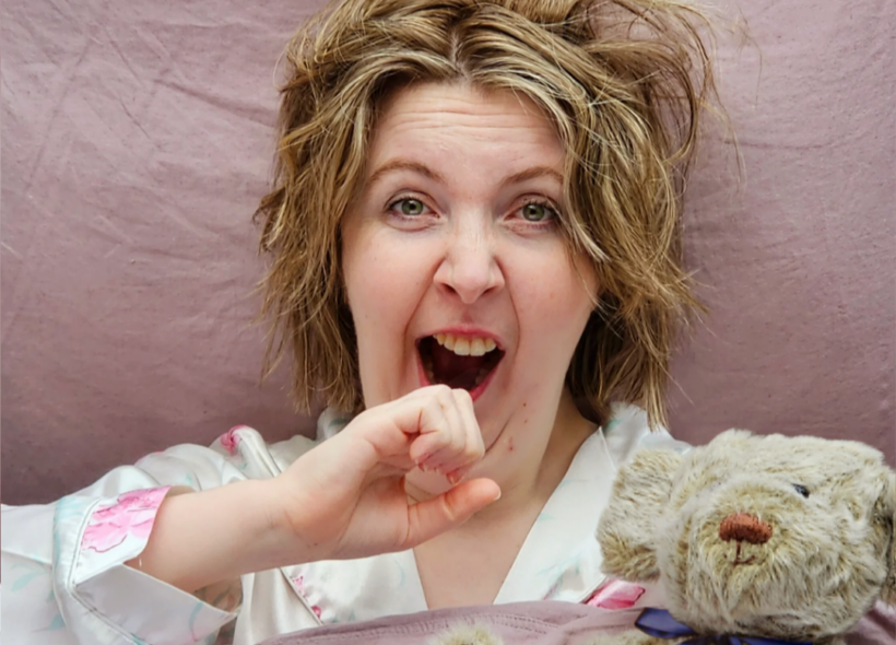 White blonde woman laying in bed yawning whilst holding teddy