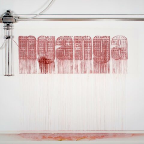 Installation view of Robert Andrews' work 'Moving Out of Muteness' 2013. Features a white wall with the word nganga written in red by a machine which is also mounted on the wall. 