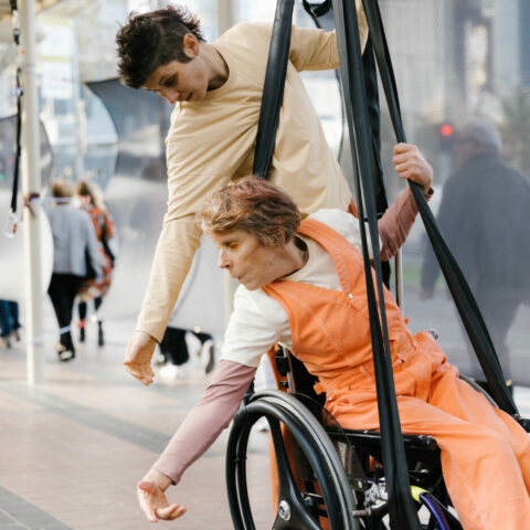 A wheelchair user is suspended in their chair above a public walkway. A fellow artist hangs from the back of the chair.