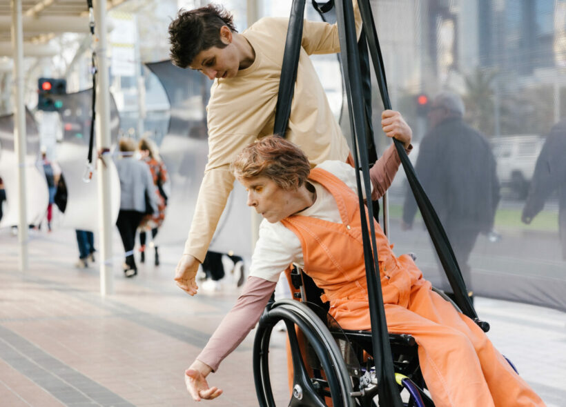 A wheelchair user is suspended in their chair above a public walkway. A fellow artist hangs from the back of the chair.