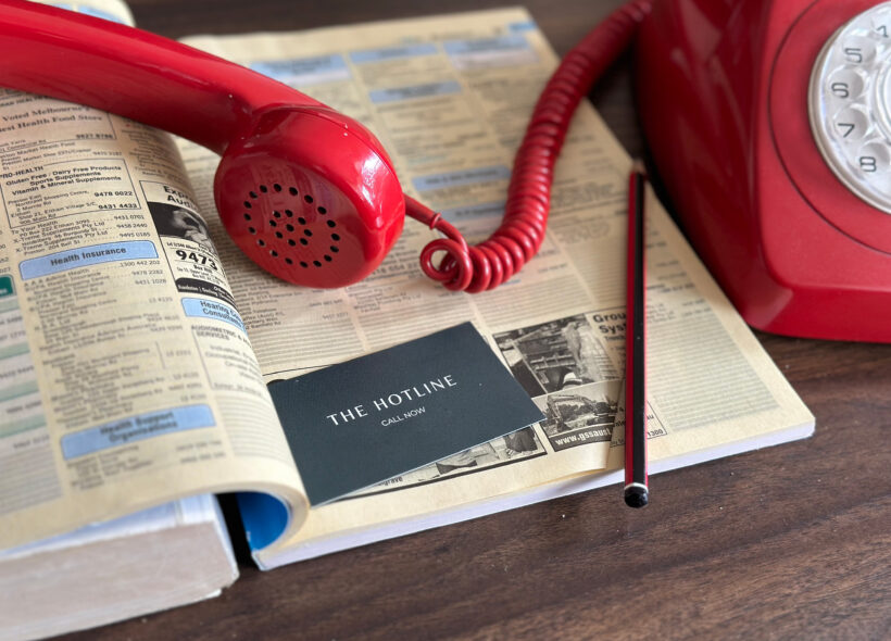 The receiver and cord for a red rotary phone lies on top of an open Yellow Pages. A pencil and a plain green business card also sits on the page. The card reads: The Hotline. Call now.