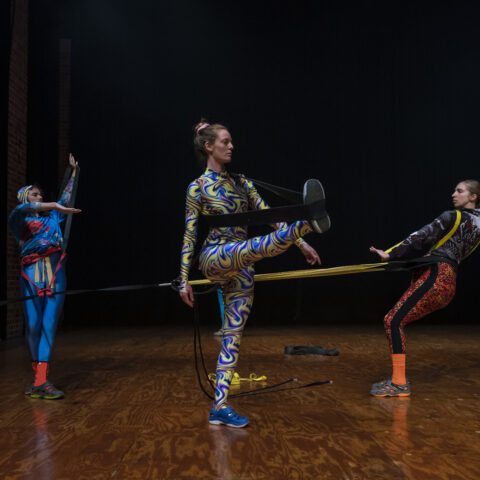 Three dancers are using exercise bands to make large movements with limbs while attached to one another.