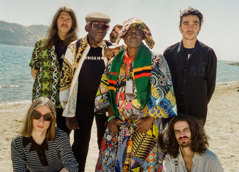 6 members of the band WITCH posing in colourful patterned clothing in front of a sunny beach backdrop. 