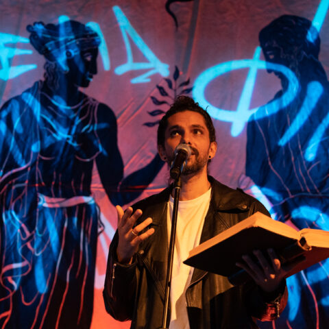 Luka Lesson performs open book in hand in front of a projection of Greek statues and Ancient Greek letters