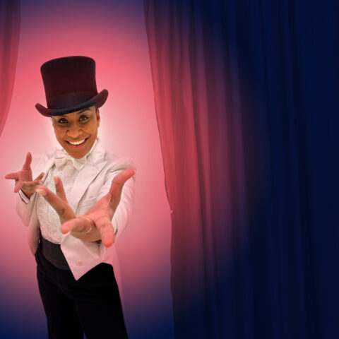 Elenoa Rokobaro, performing as Nellie Small, stands in a suit (white blazer, black pants and a top hat) in front of red curtains, arms stretched out forward