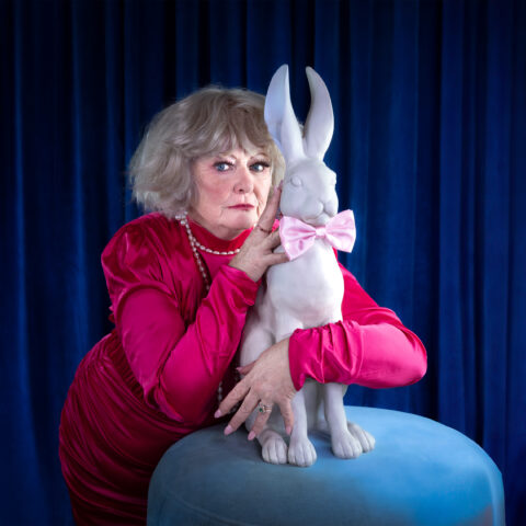A blue curtain background. A performer wears a pink satin blouse and a pearl  necklace. They make eye contact with the camera whilst hugging a white rabbit that is on a light blue table. The rabbit has a baby pink bow around its neck.