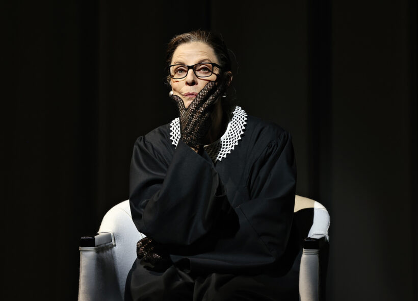 Heather Mitchell. as RBG, sits in judges robes with her chin resting on her hand. 