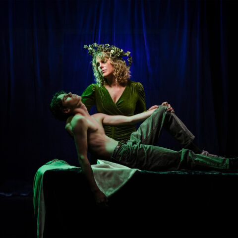 A blue curtain background that appears darker than it is. A performer wears a green velvet long sleeved dress and a wreath of olives and leaves. They cradle another performer below them who is half on a table with a white tablecloth. This performer has a bare chest and wears jeans.