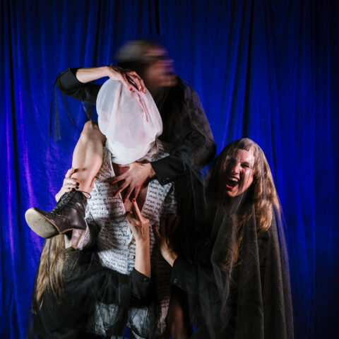 A blue curtain background. There are four figures in this image. Three figures are in black with translucent black veils. In the middle of the image, a performer is wearing a white suit with repeated black text stating “Smash the Patriarchy”. The head of the performer is covered in white cloth. The three figures in black hold onto the central figure forcing them there. One of the black figures is on the shoulders of the figure in the middle. The figure on the left has their rude finger pointing at the audience whilst the figure on the left screams.
