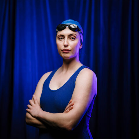 A blue curtain background. A performer stares at the camera with a bemused expression on their face. They wear a blue, one piece swimming outfit. On their head is a blue swimming cap and goggles. Their arms are crossed. 