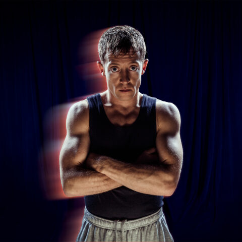 A blue curtain background that appears almost black. A performer stands with their arms crossed. They wear a navy blue muscle tee and gray tracksuit pants. The camera is used with a slow shutter speed to give the illusion of movement. 