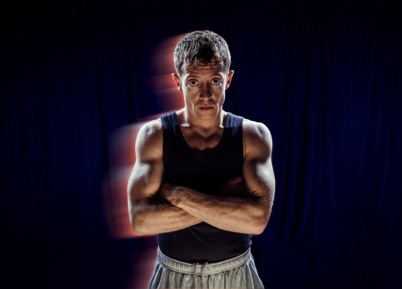 A blue curtain background that appears almost black. A performer stands with their arms crossed. They wear a navy blue muscle tee and gray tracksuit pants. The camera is used with a slow shutter speed to give the illusion of movement. 