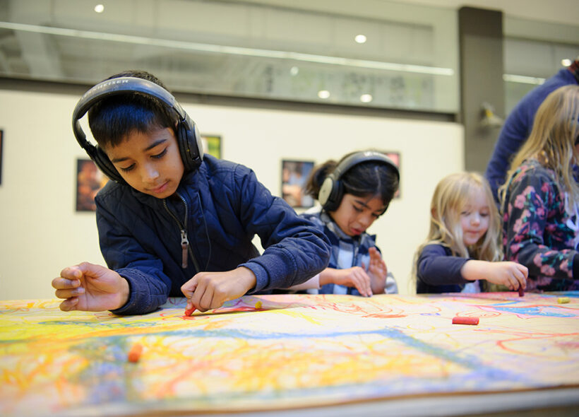 A Sound of Drawing production photo. A child wearing a jacket and headphones, draws intently on a table covered in paper. Other children are visible behind them, also drawing. Photo: Sarah Walker