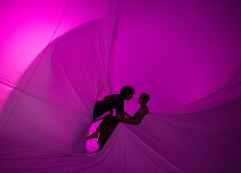 A silhouette of a man and woman is at the centre of the image. The woman is sitting down while the man emerges from an inflatable leaning into her. They are surrounded by a magenta-coloured fabric.