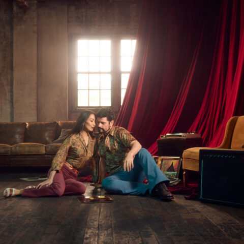 A man & a woman lean towards each other. They are dressed in a bohemian style, in a semi-bare warehouse apartment