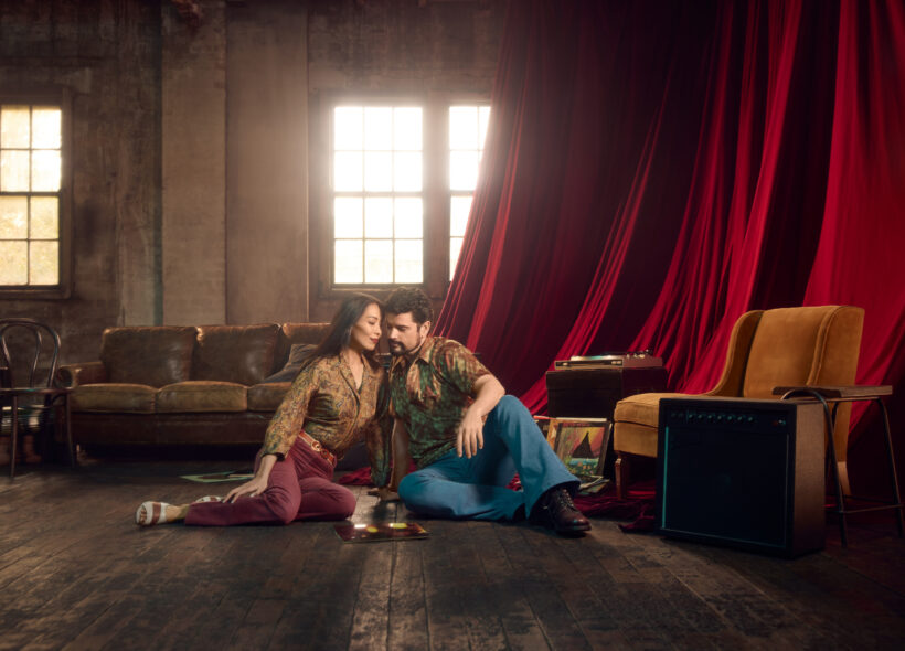 A man & a woman lean towards each other. They are dressed in a bohemian style, in a semi-bare warehouse apartment