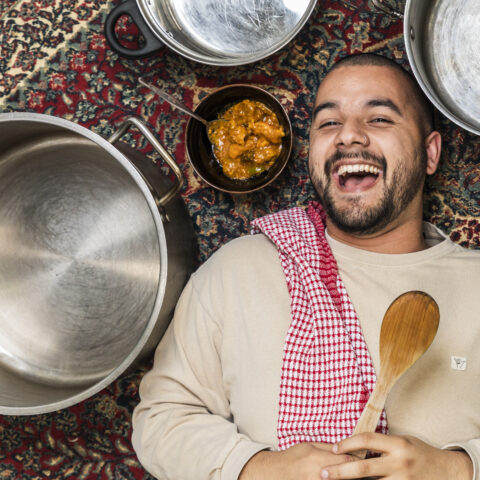 Joshua Hinton lays on a persian rug surrounded by empty cooking pots and a bowl of curry. He laughs with a red tea towel over his shoulder and holds a spoon. 