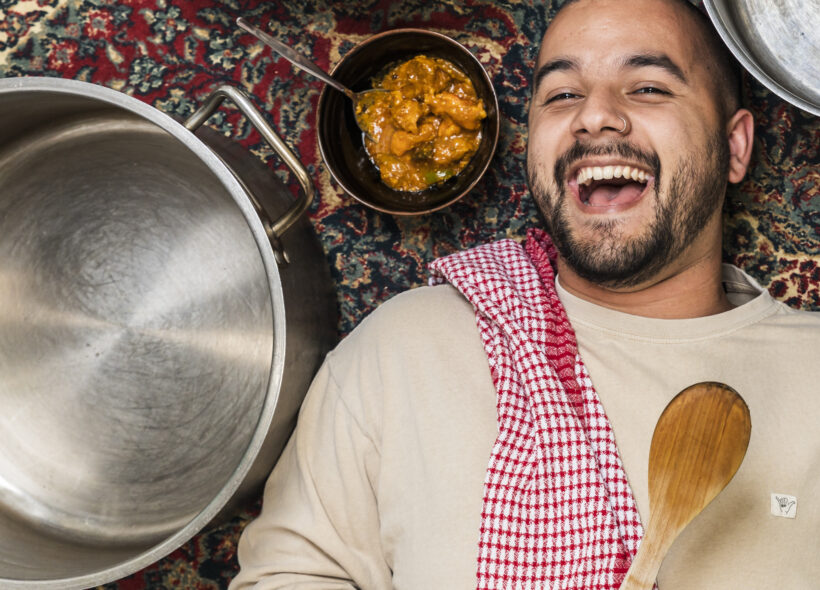 Joshua Hinton lays on a persian rug surrounded by empty cooking pots and a bowl of curry. He laughs with a red tea towel over his shoulder and holds a spoon. 