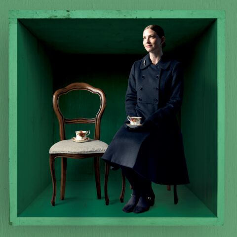 Elizabeth Blackmore sits in a black overcoat with a tea cup in hand, next to an empty chair with a teacup on it. The background is green.