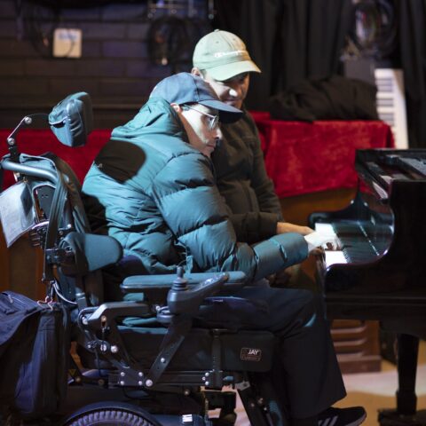 A man in his wheelchair plays piano with his support worker sitting by his side