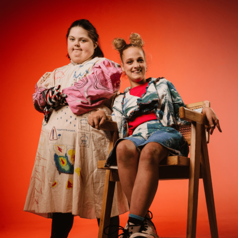 Two women dressed in handmade fashion creations look at the camera smiling; one standing with arms crossed, one sitting with legs crossed. Red backdrop. 