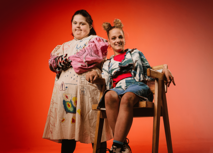 Two women dressed in handmade fashion creations look at the camera smiling; one standing with arms crossed, one sitting with legs crossed. Red backdrop. 