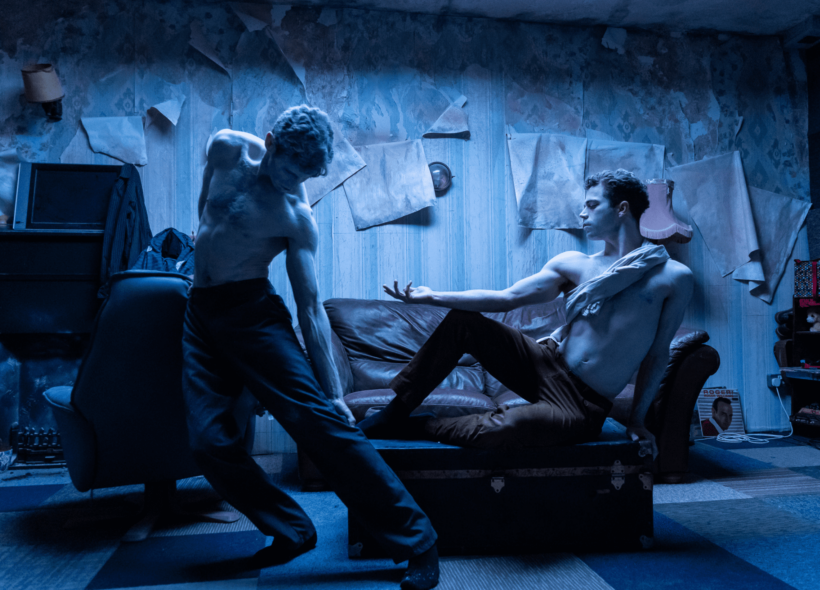 Two shirtless male dance actors in trousers mid-dance pose; left dancer stands bending with left arm extended and resting on left leg, right dancer reclines on a leather two seater couch with right knee at 90 degrees and right extended arm resting atop - set in a loungeroom with with peeling wallpaper and the whole image is overlaid with an eerie navy blue light