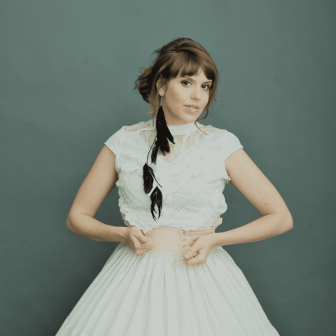 Performer Pauline Maudy in a white lace short-sleeve top and white skirt with one long black feather earring standing in front of a grey backdrop