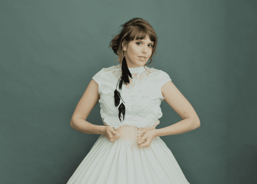 Performer Pauline Maudy in a white lace short-sleeve top and white skirt with one long black feather earring standing in front of a grey backdrop