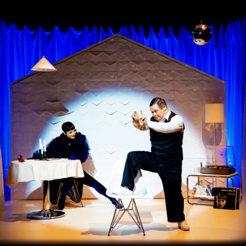 Still image of live theatre scene: Blue velvet curtain backdrop, male actor to right in foreground with leg on white chair with arms raised, female actor to left in background wearing feather boa and red lipstick at dining table. Record player and lamp in background right corner.