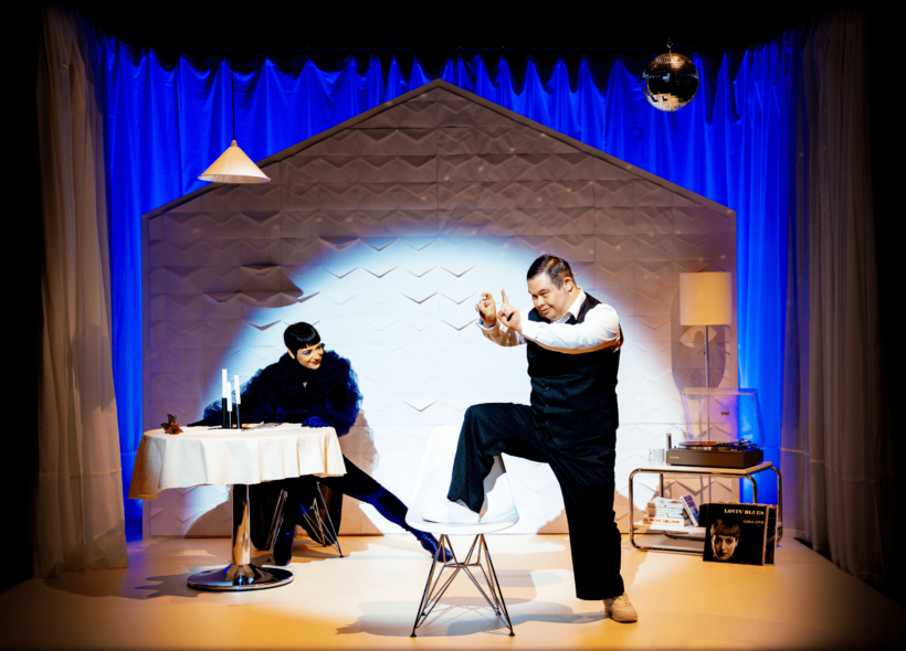 Still image of live theatre scene: Blue velvet curtain backdrop, male actor to right in foreground with leg on white chair with arms raised, female actor to left in background wearing feather boa and red lipstick at dining table. Record player and lamp in background right corner.