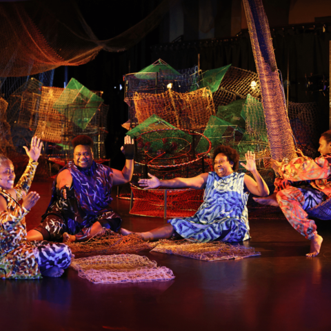 Four Torres Strait Islander women dressed sit on stage in a semi circle around weaving wares, in the middle of song, hands clapping and smiling. The stage is draped in colourful nets and crabpots and fishtraps and backlit with warm lighting.