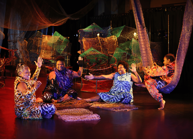 Four Torres Strait Islander women dressed sit on stage in a semi circle around weaving wares, in the middle of song, hands clapping and smiling. The stage is draped in colourful nets and crabpots and fishtraps and backlit with warm lighting.