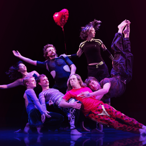 A group of 7 dance actors form a human sculpture mid dance, leaning on one another in different poses on a dark stage, a red heart helium balloon hangs in the air