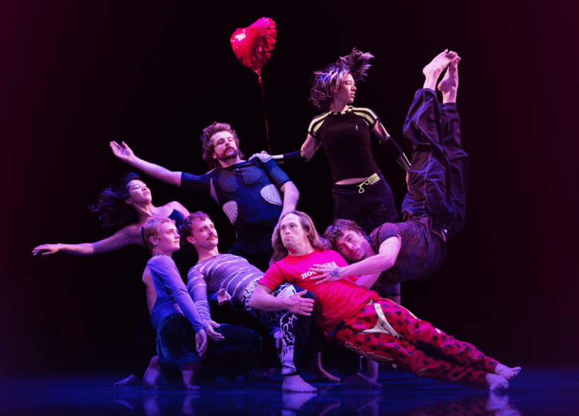 A group of 7 dance actors form a human sculpture mid dance, leaning on one another in different poses on a dark stage, a red heart helium balloon hangs in the air