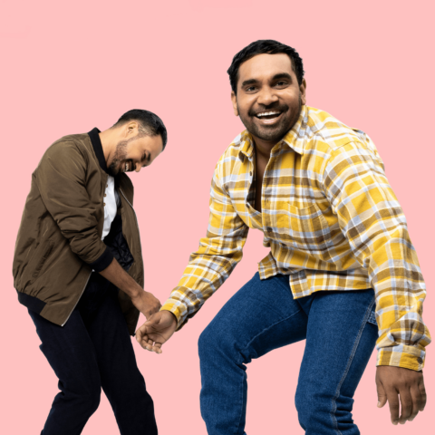 Two young Indigenous (Murri) men smiling: one in foreground in yellow plaid shirt stands smiling directly at the camera, one in background in brown jacket stands smiling head down - pink background