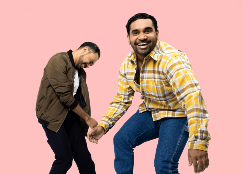 Two young Indigenous (Murri) men smiling: one in foreground in yellow plaid shirt stands smiling directly at the camera, one in background in brown jacket stands smiling head down - pink background