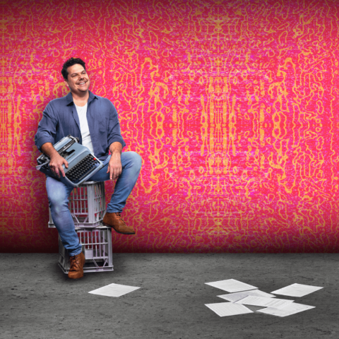 Actor sitting on two milk crates, smiling and holding typewriter, paper scattered on floor, pink backdrop