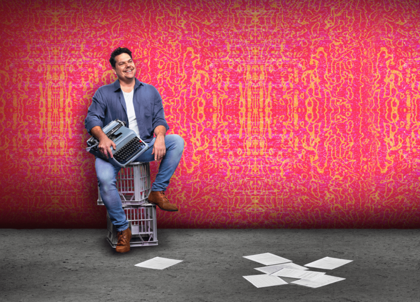 Actor sitting on two milk crates, smiling and holding typewriter, paper scattered on floor, pink backdrop