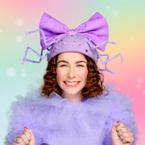 Main actress Fancy Long Legs smiles excitedly at the camera, wearing a purple cap and bow and purple sparkly costume in front of a rainbow backdrop