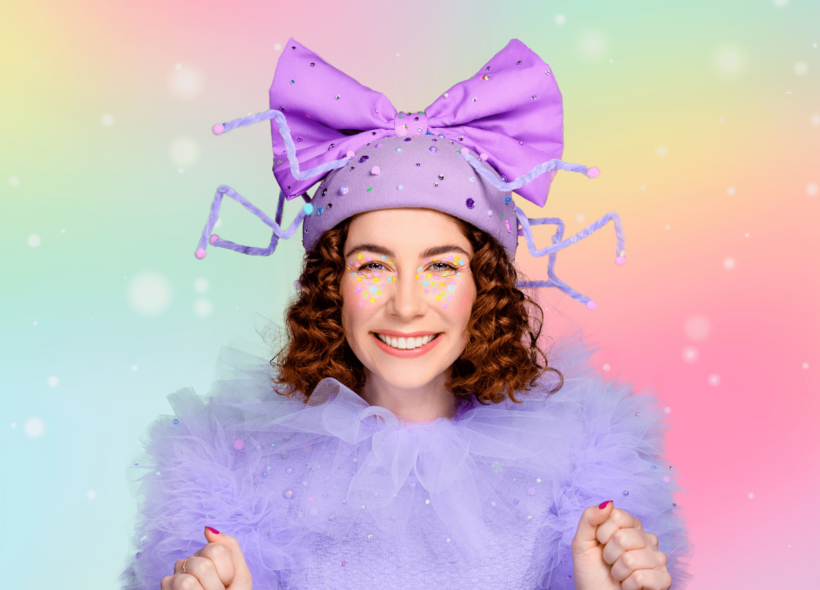 Main actress Fancy Long Legs smiles excitedly at the camera, wearing a purple cap and bow and purple sparkly costume in front of a rainbow backdrop